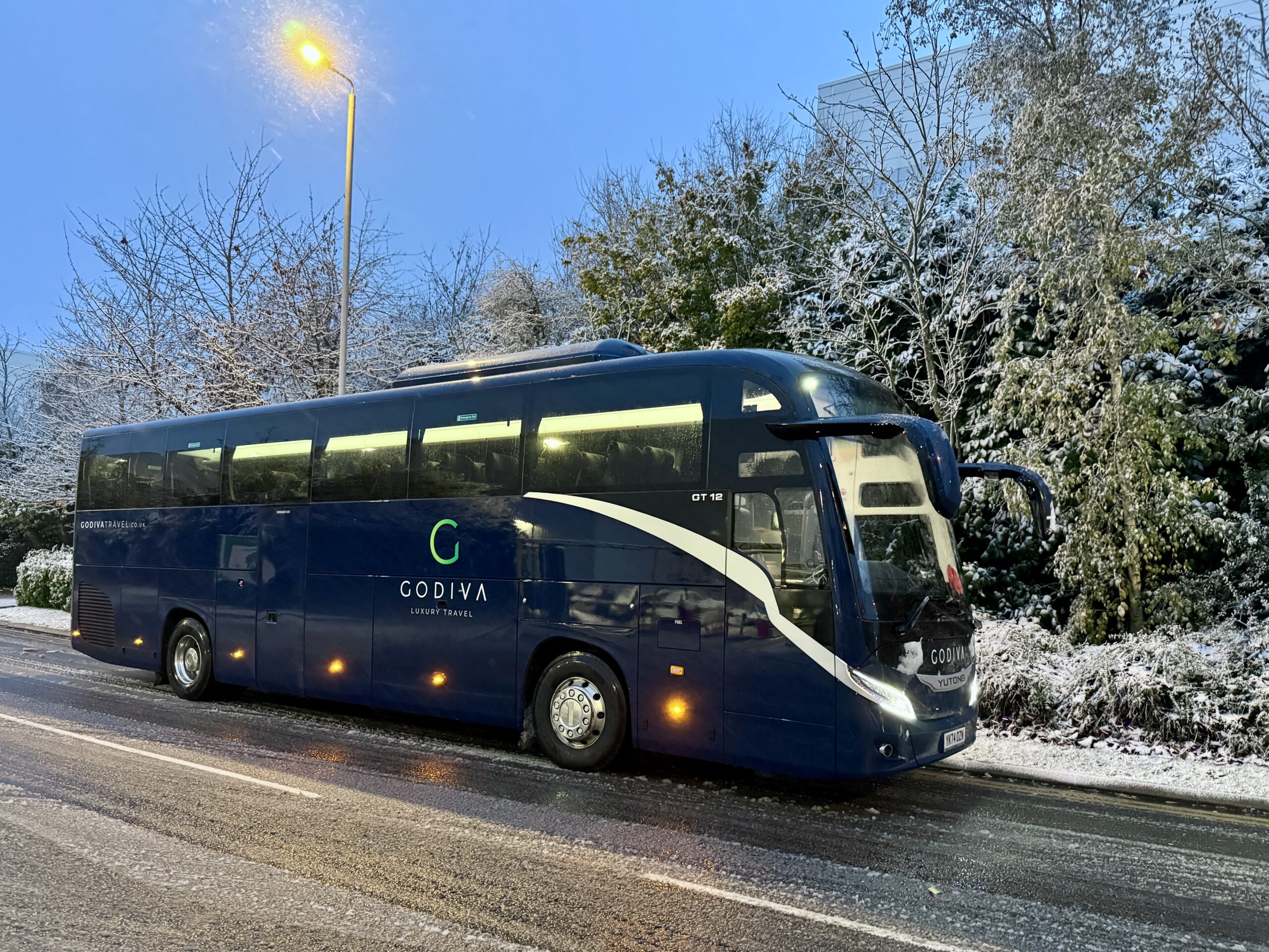 A blue Godiva Travel Yutong coach parked next to trees surrounded by winter snow.